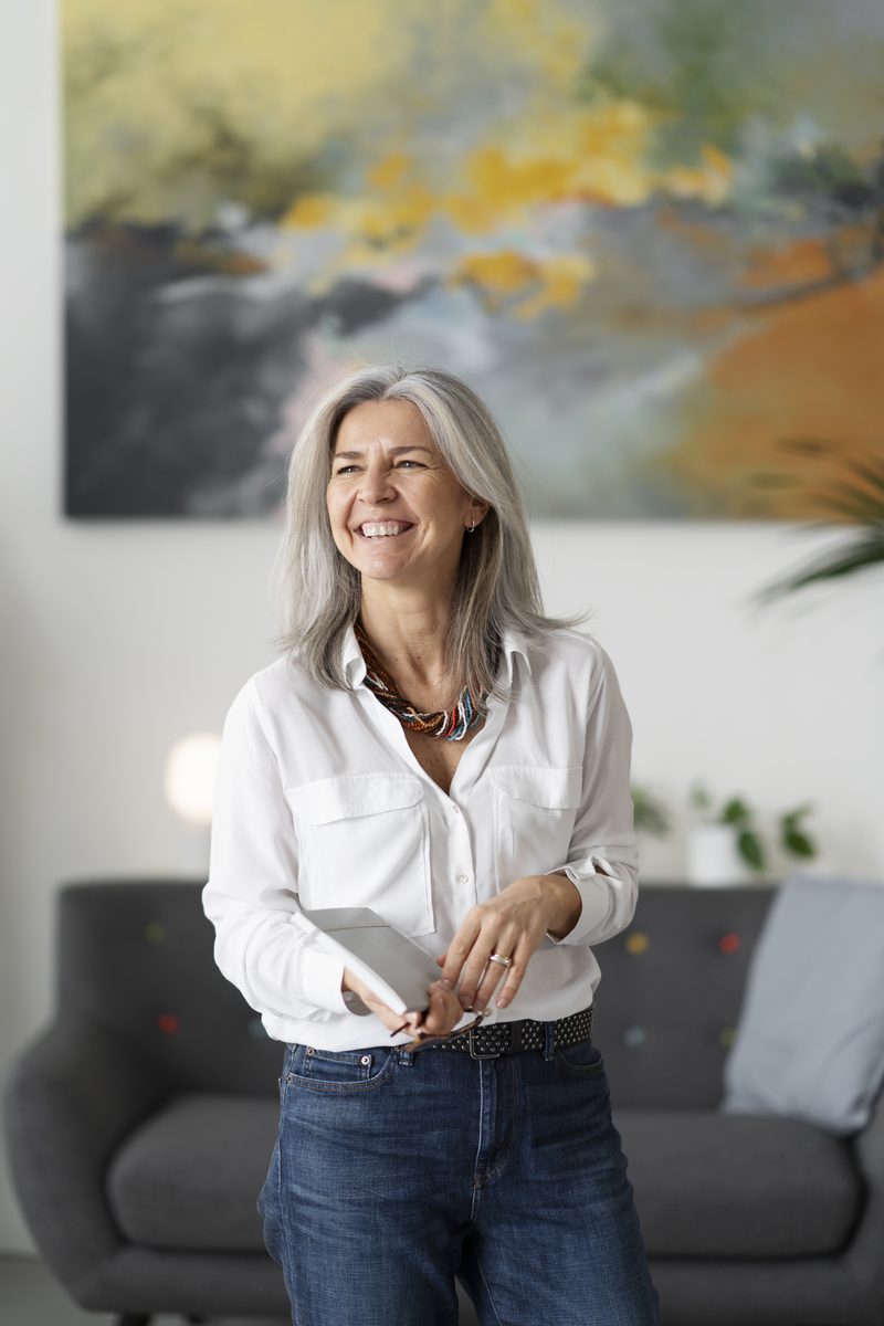 A woman with grey hair in a white shirt, smiling, standing in a bright room